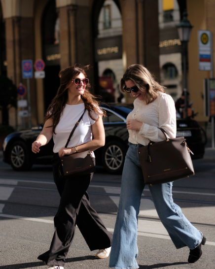 Two women walking on a city street, each carrying a Vienna handbag. The woman on the left wears a white top and black wide-leg pants, while the woman on the right wears a cream textured top and light blue wide-leg jeans.