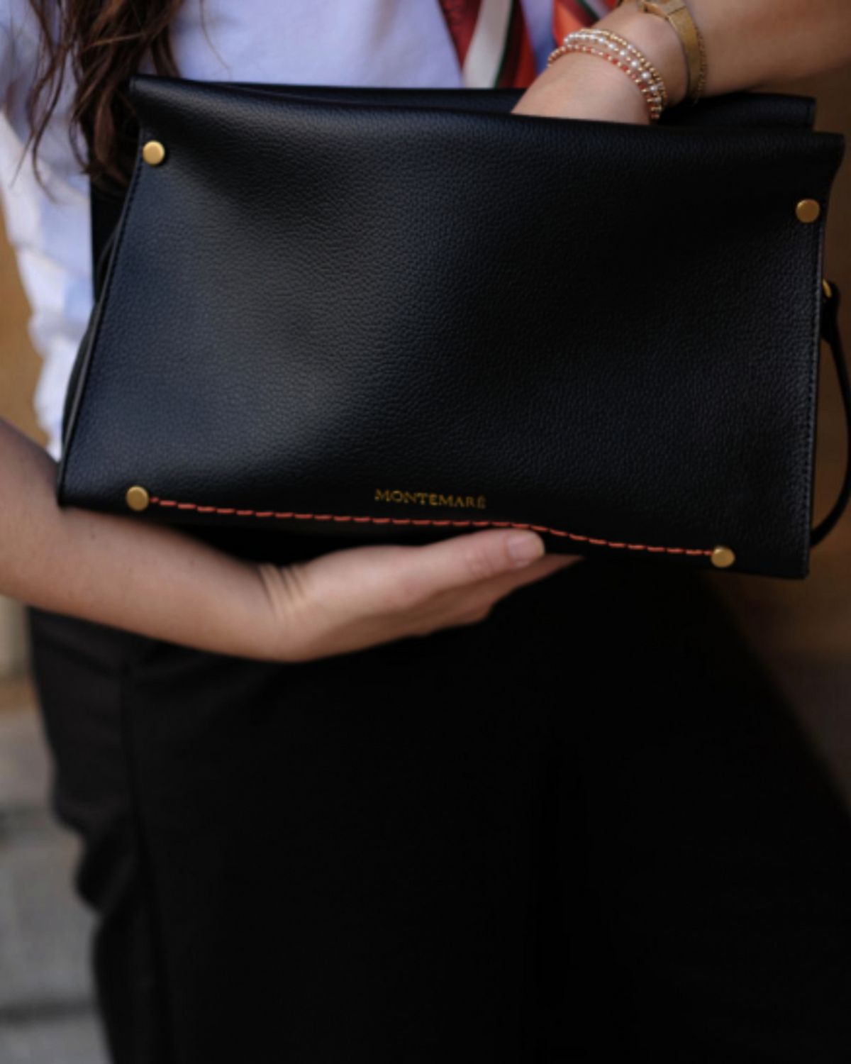 A person holding a Montemare Small Shoulder Bag in black with gold-tone hardware and orange contrast stitching.