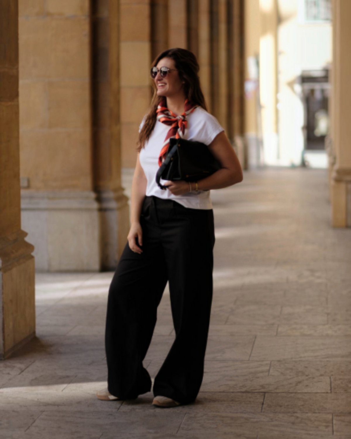 A woman wearing white shirt, black pants, and Montemare's Silk Scarf carrying a black Vienna Small Shoulder Bag.