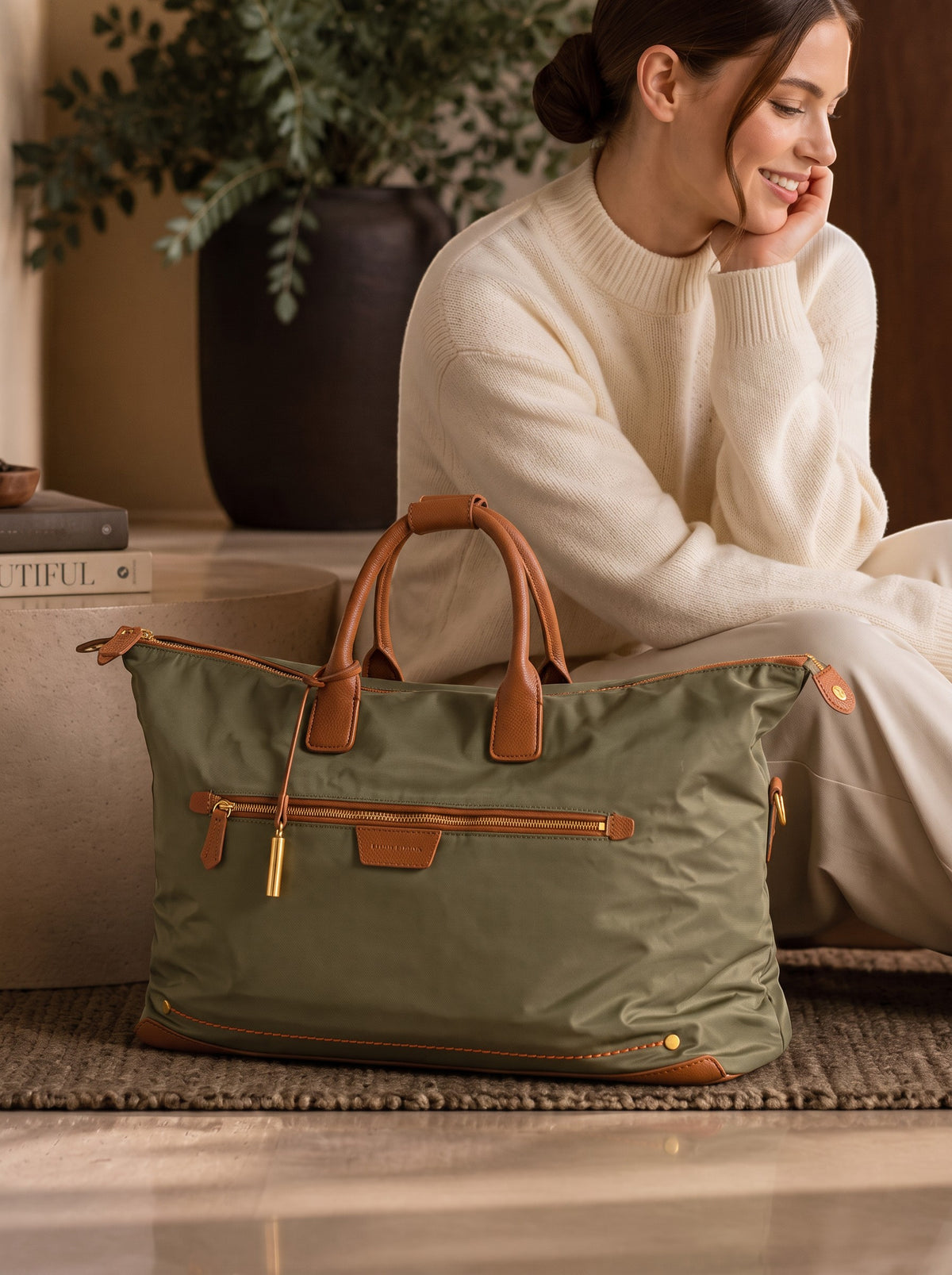 An olive green Oslo Carry-On bag with tan leather handles and trim sits on a rug in a styled indoor setting, with a woman in a cream sweater sitting behind it.