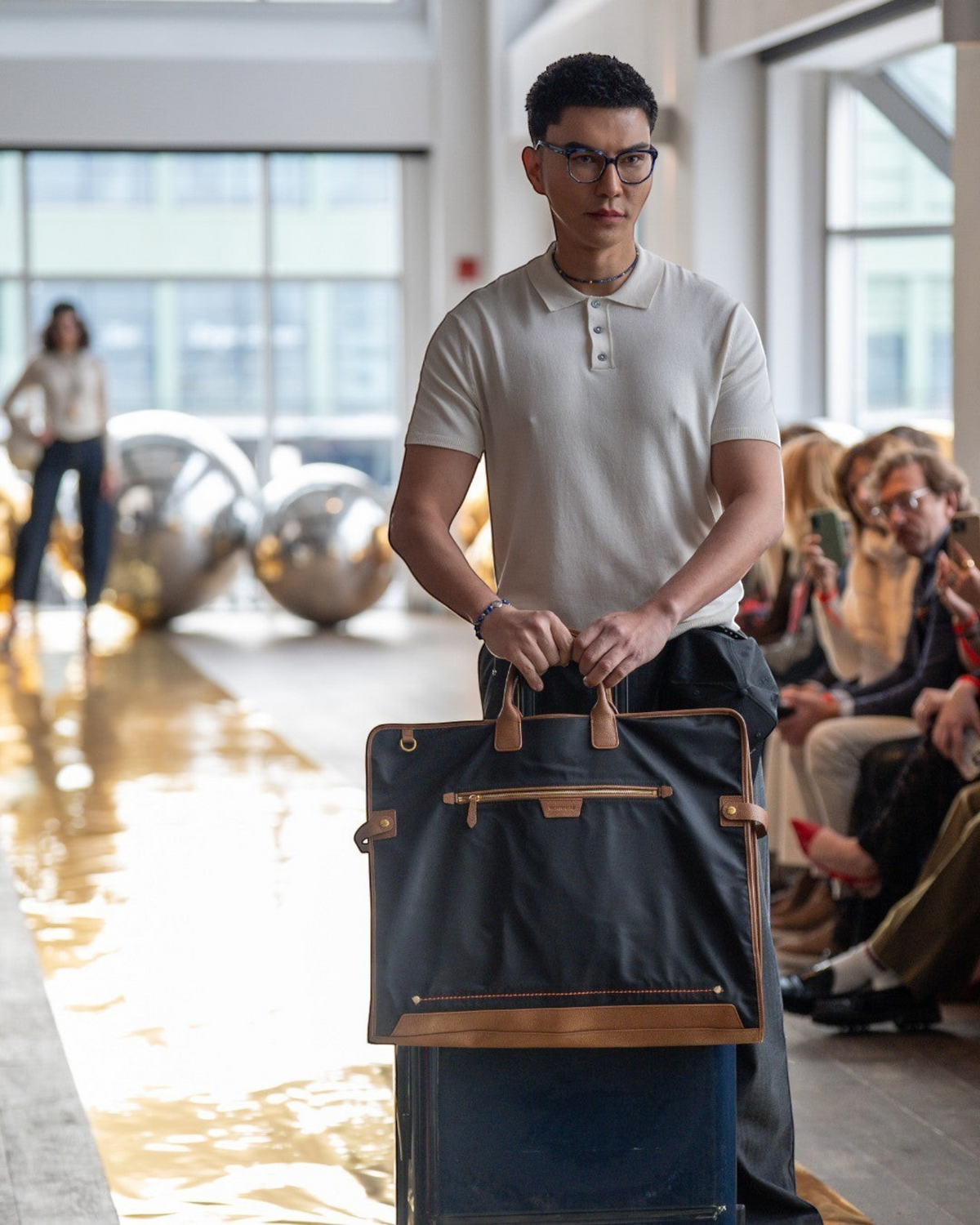 A model carries a black Montemaré tri-fold garment bag with leather trim on a runway during a fashion show.