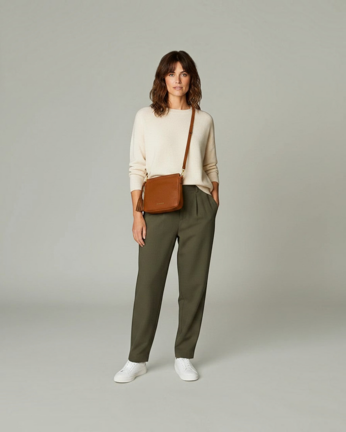 A woman wearing a Montemaré brown leather square pouch crossbody bag with a tassel detail over a cream sweater.