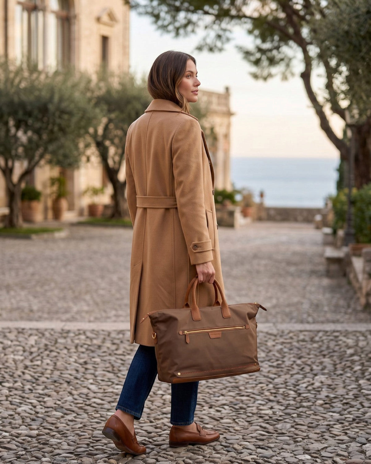 Woman carrying a brown Montemaré Oslo waterproof nylon and leather weekender bag on a cobblestone street.