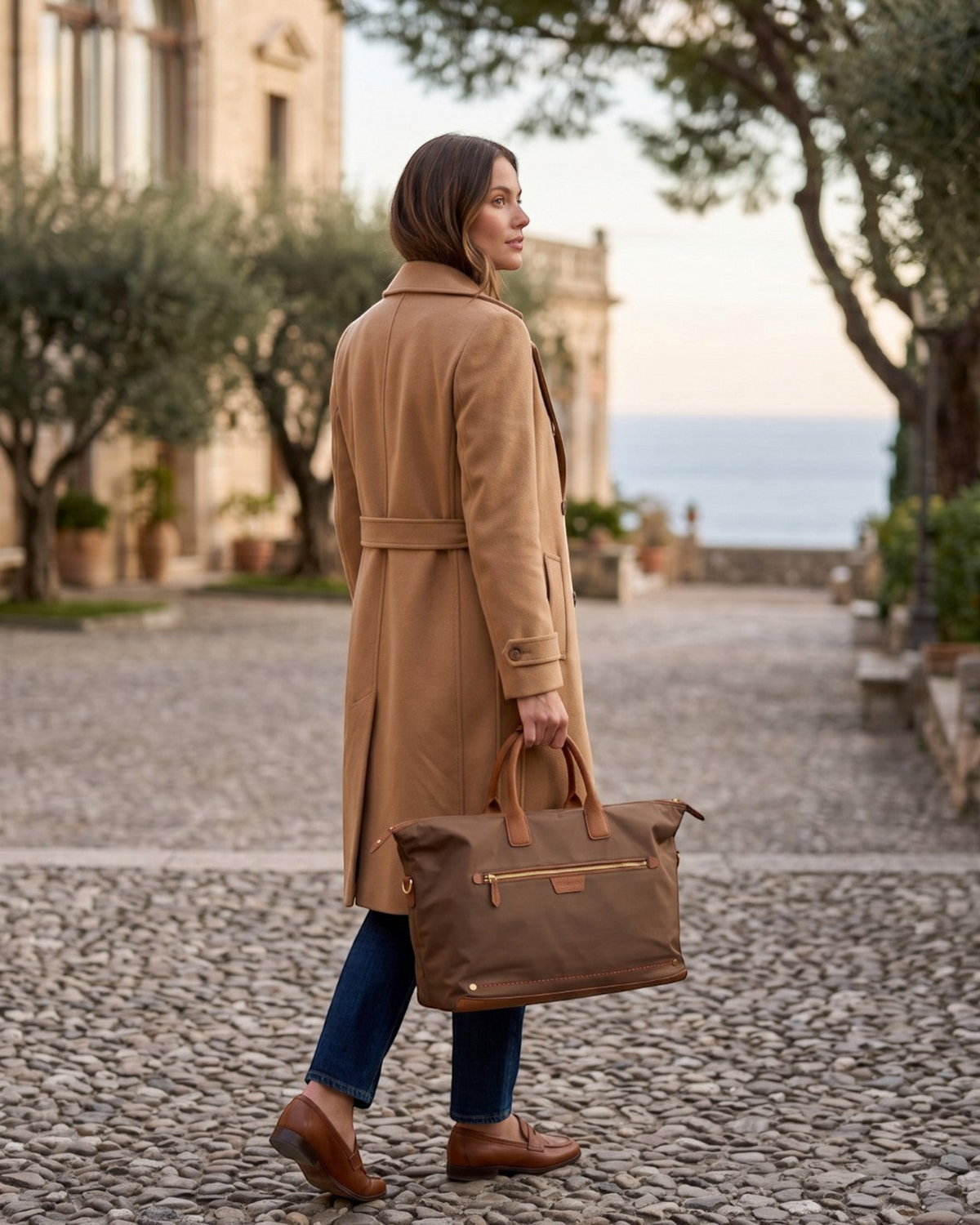 Woman carrying a brown Montemaré Oslo waterproof nylon and leather weekender bag on a cobblestone street.