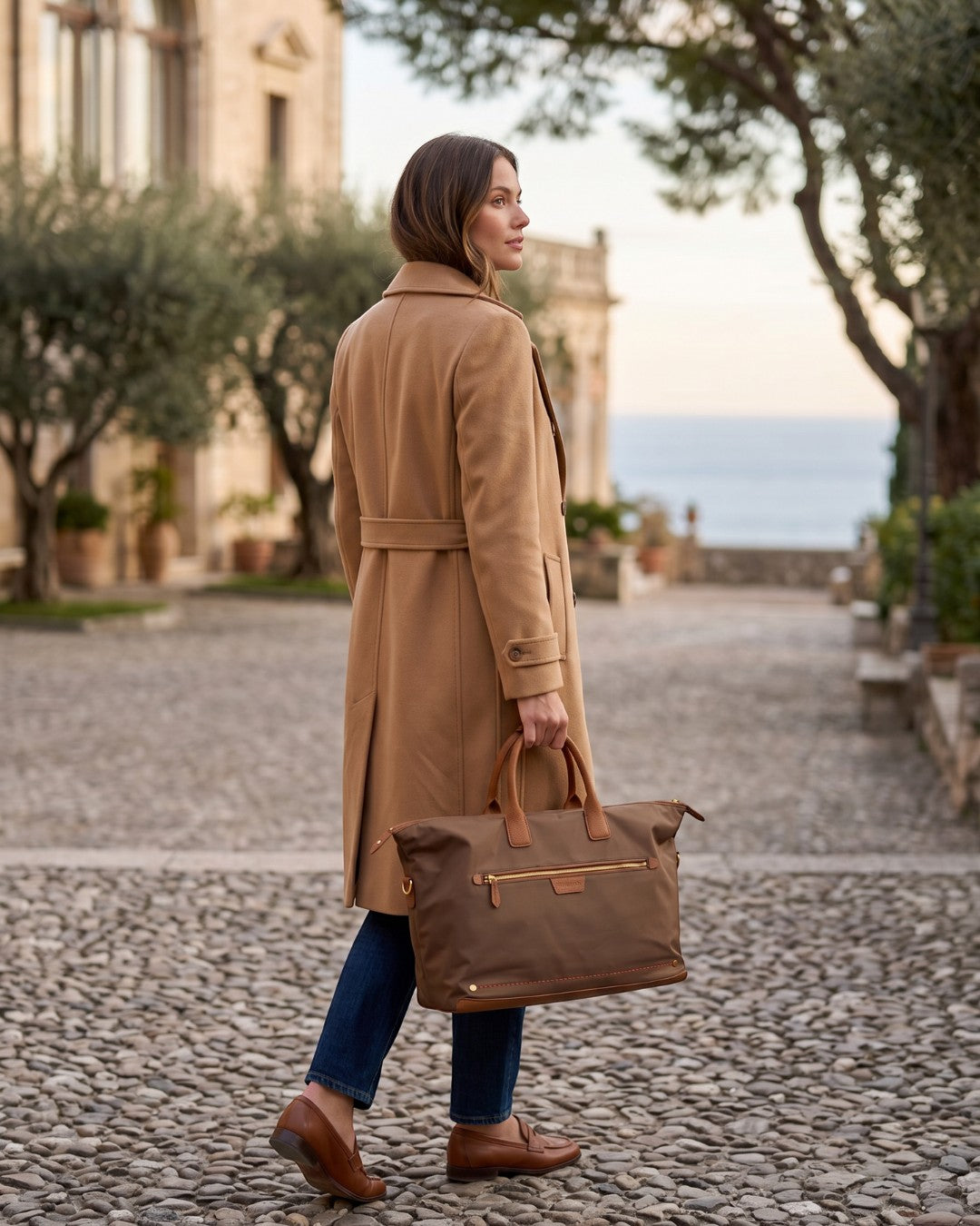 Montemaré Oslo Carry On weekender bag in brown waterproof nylon with leather trim held by a woman in a camel coat.