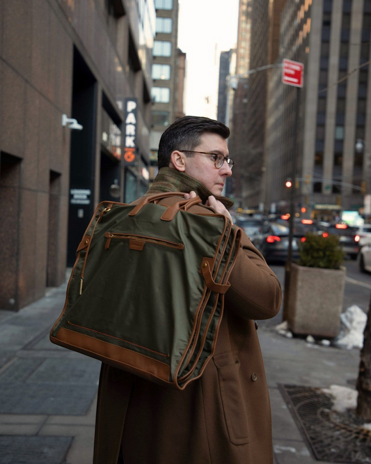 Man carrying a Montemaré Oslo tri-fold garment bag in olive waterproof nylon with brown leather trim on a city street.