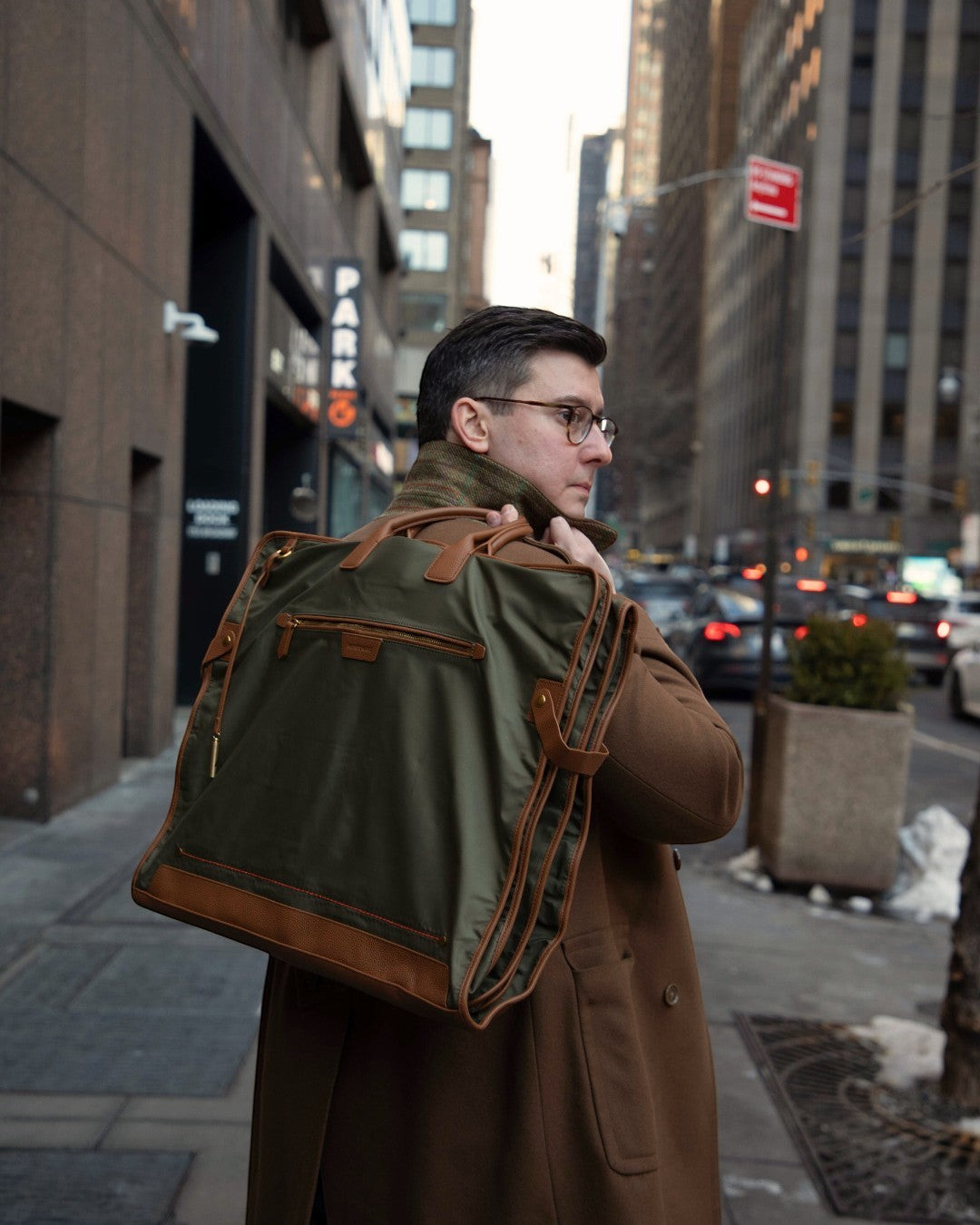 Man carrying a Montemaré Oslo tri-fold garment bag in olive waterproof nylon with brown leather trim on a city street.