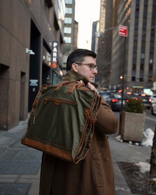 Man carrying a Montemaré Oslo tri-fold garment bag in olive waterproof nylon with brown leather trim on a city street.