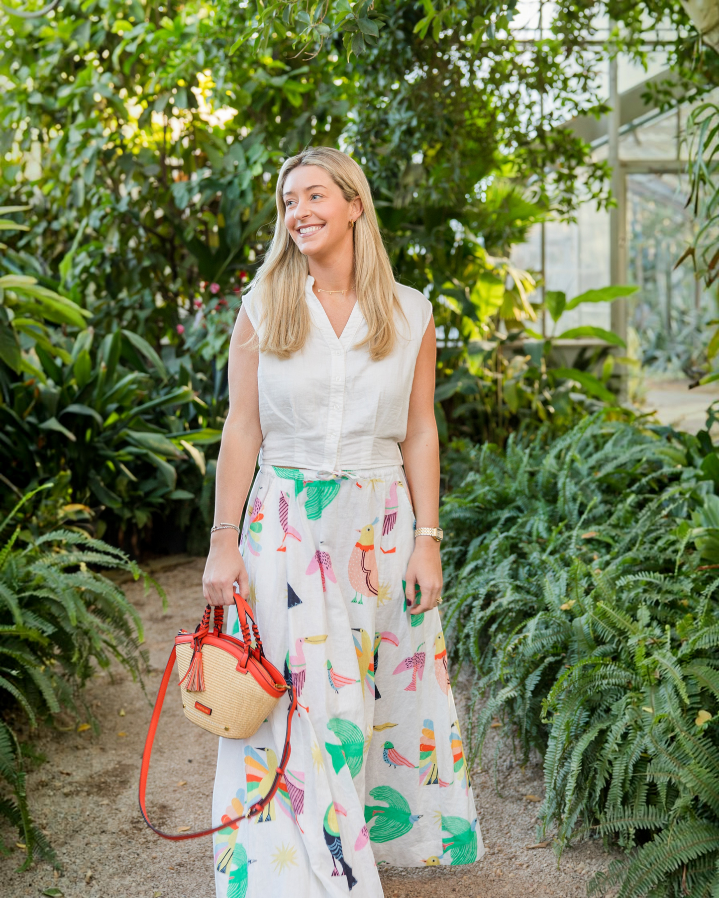 Woman carrying a Montemaré Palma straw-style crossbody bag with orange vegan leather trim in a lush greenhouse
