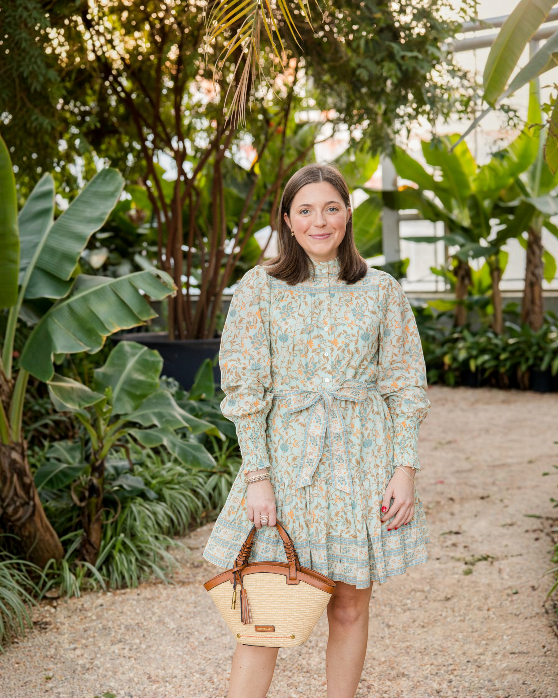 Woman wearing a floral dress holding a Montemaré Palma straw-style crossbody bag with vegan leather trim and tassel