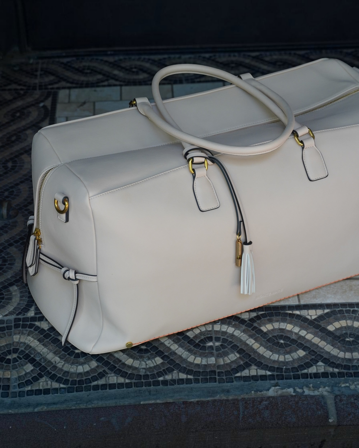 Montemaré Roma vegan leather duffel bag in cream with gold hardware, tassel detail, and orange interior trim.