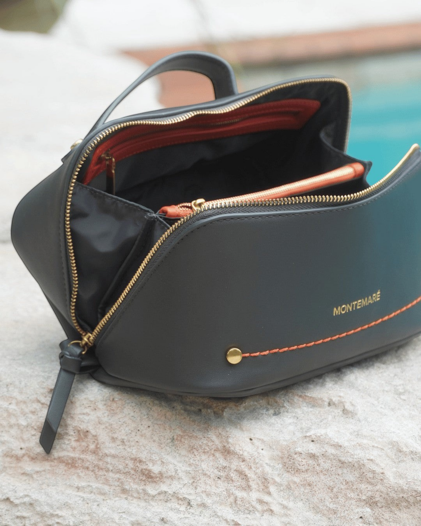 Montemaré Roma foldout toiletry case in black vegan leather with orange stitching and gold hardware on stone surface