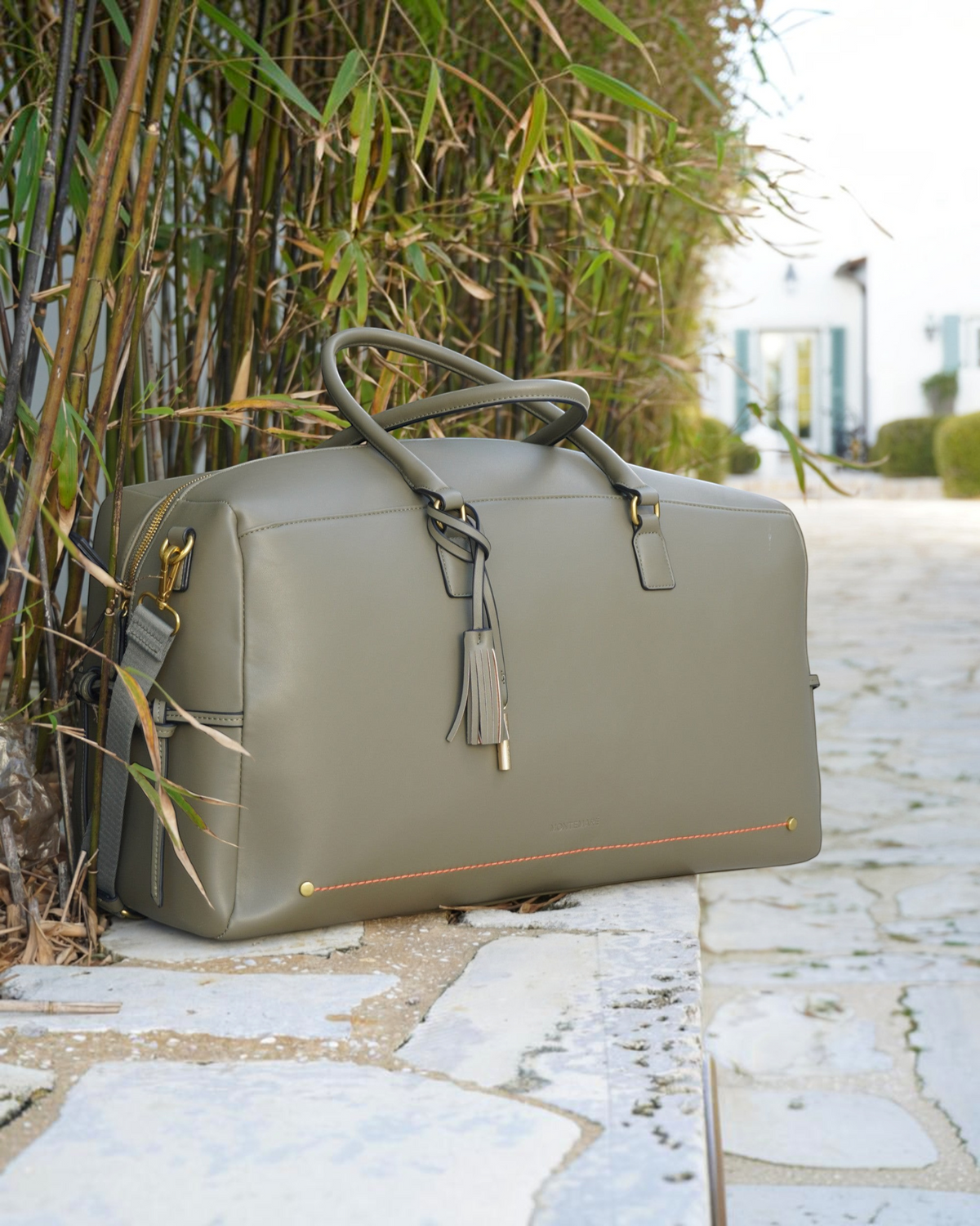 Montemaré Roma olive green vegan leather travel duffel bag with a tassel and gold hardware on a stone wall.