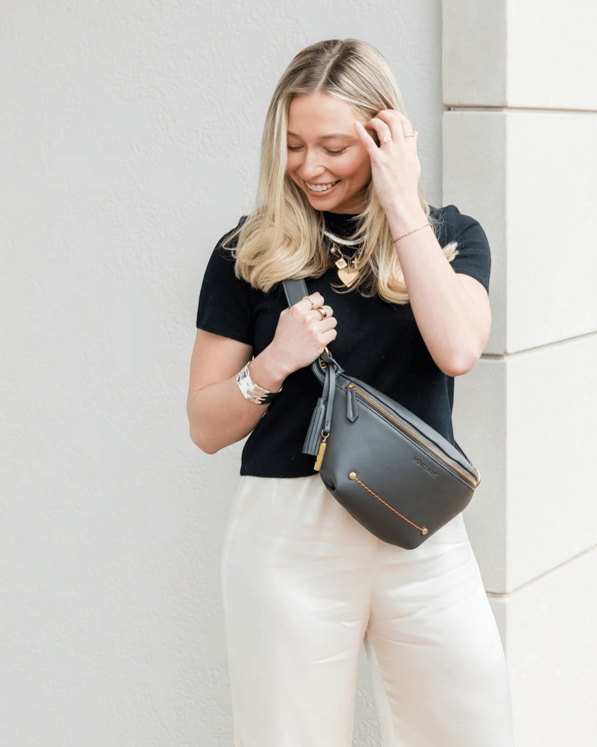 Montemaré Roma Sling bag in charcoal vegan leather worn crossbody by a smiling woman in a black top and white pants