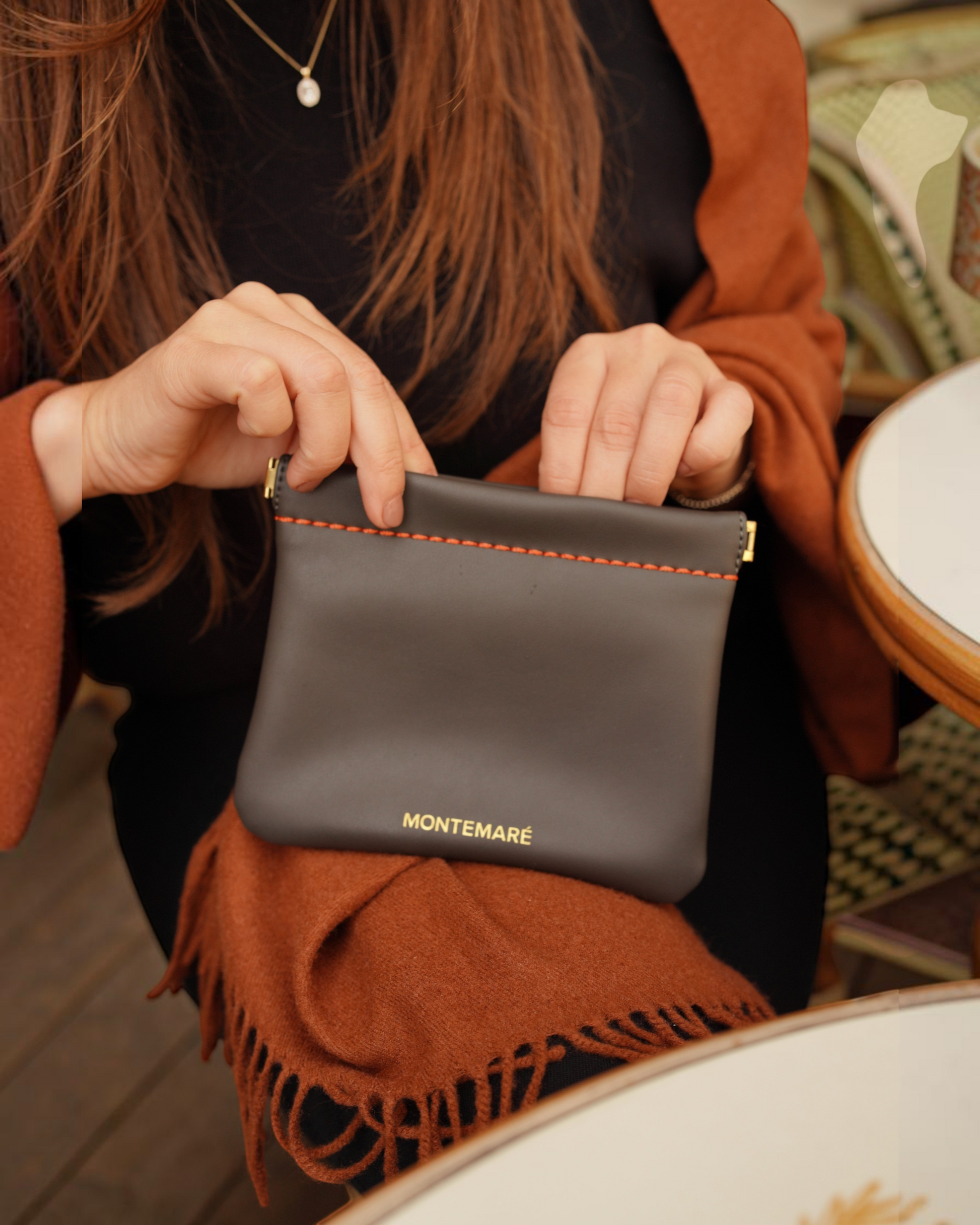 Montemaré Roma square pouch in dark grey vegan leather with orange stitching, held by a woman at a cafe table.