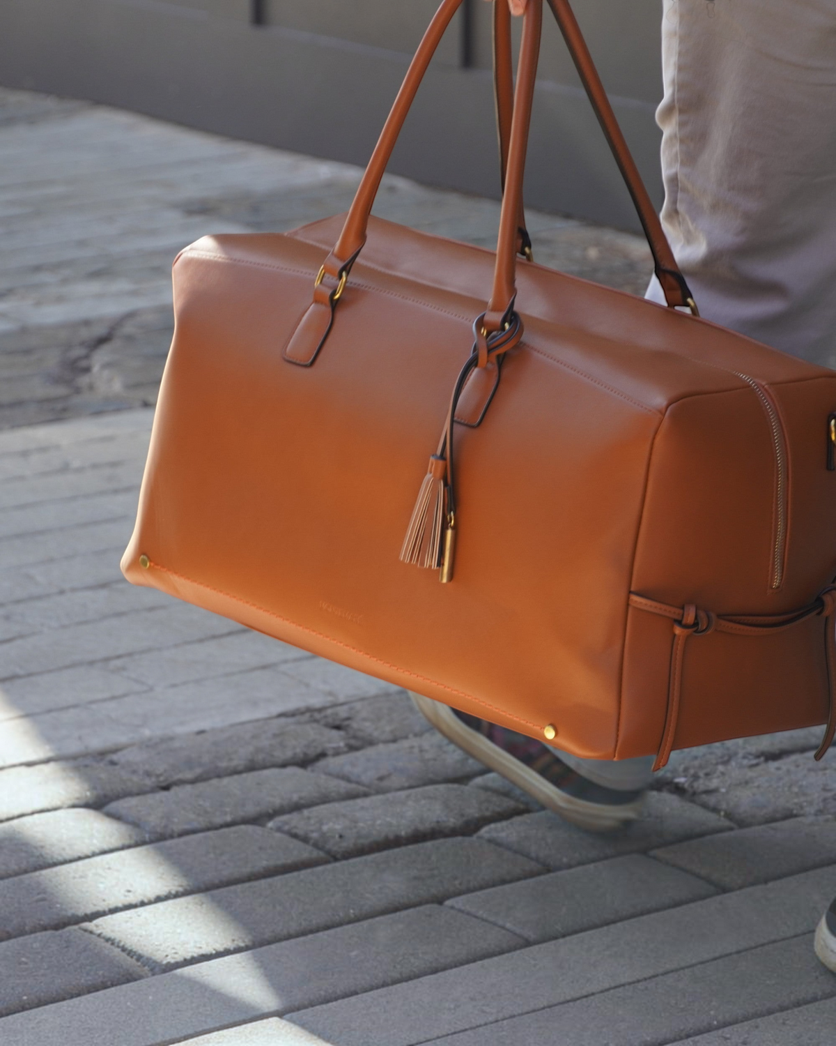 Montemaré Roma tan vegan leather travel duffel bag with a tassel detail, carried by a person on a stone walkway.