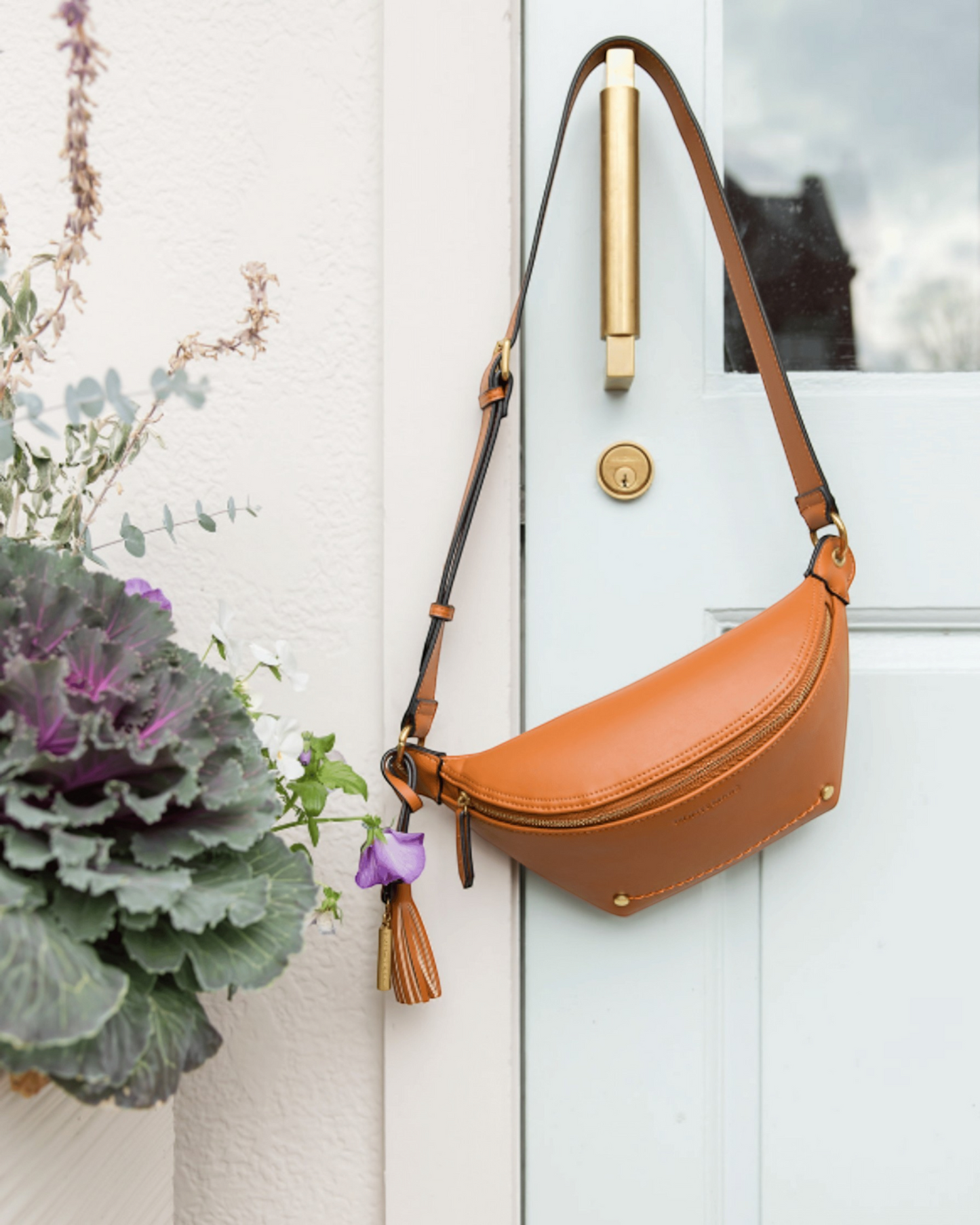 Montemaré Roma tan vegan leather sling bag with adjustable strap and signature safety whistle tassel hanging on a door