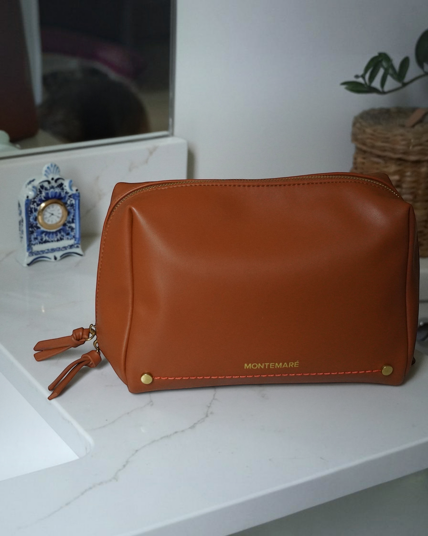 Montemaré Roma standing toiletry case in tan vegan leather with gold hardware, sitting on a white bathroom counter.
