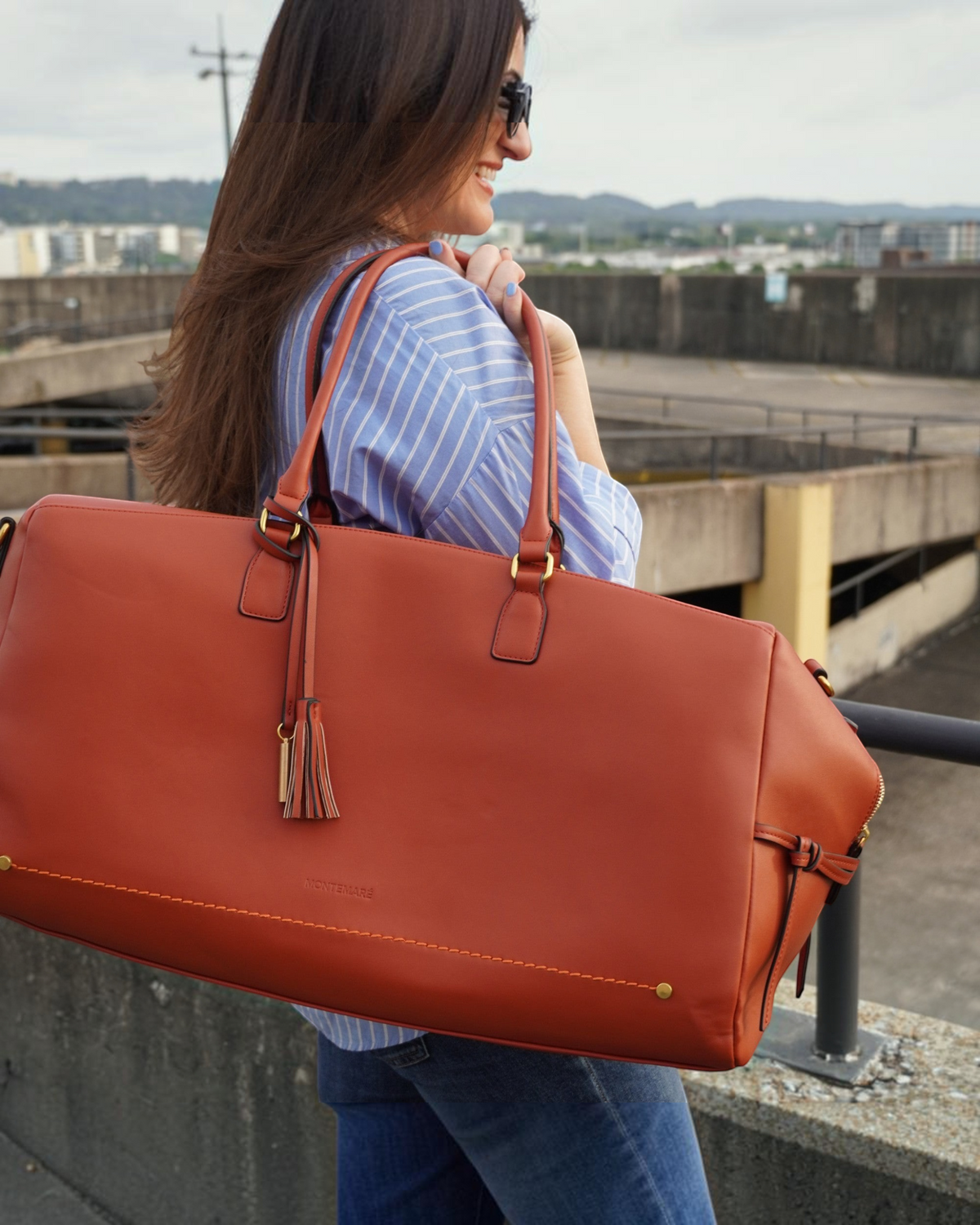 Woman carrying a tan Montemaré Roma vegan leather duffel bag with a tassel detail over her shoulder outdoors.