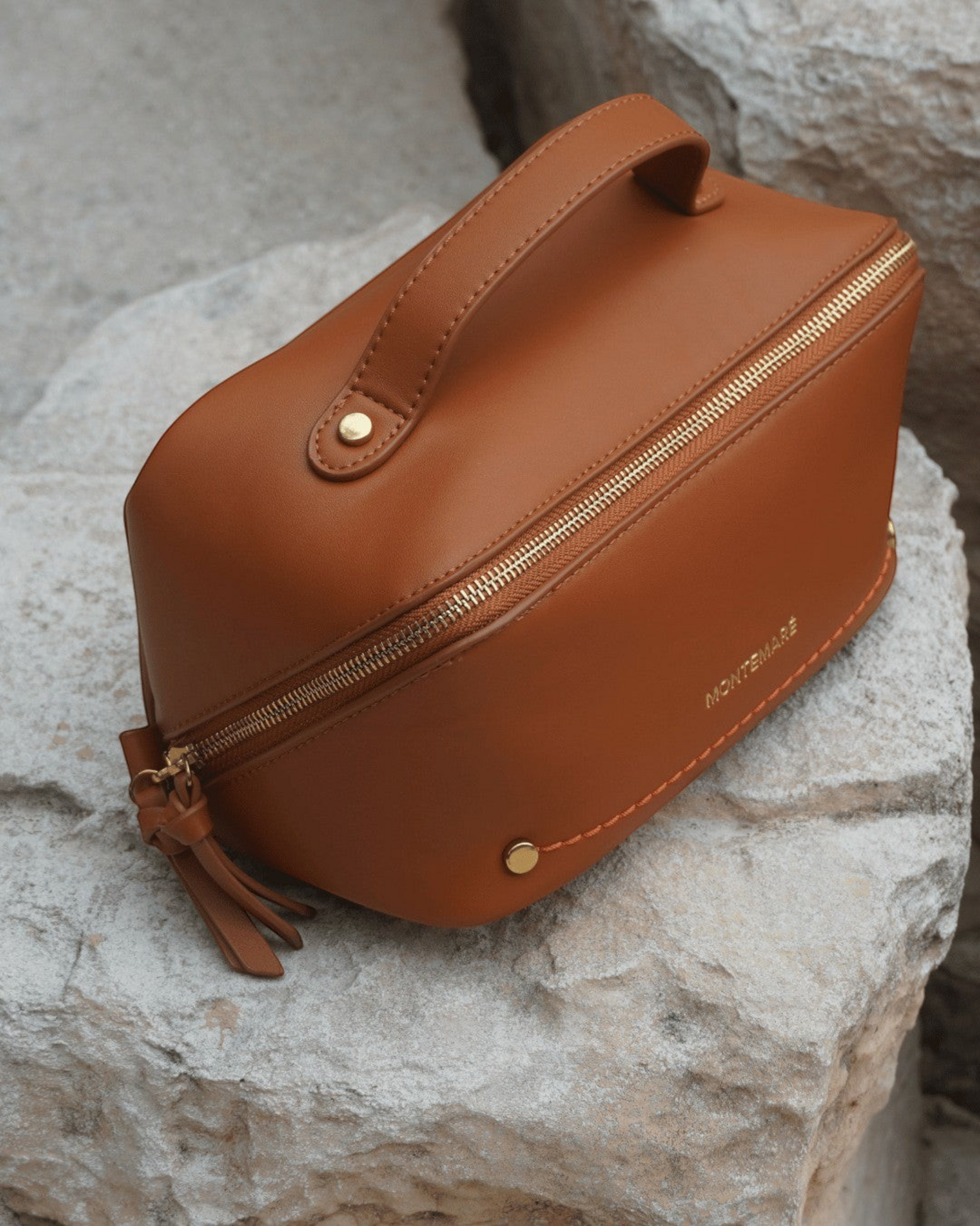 Montemaré Roma foldout toiletry case in orange vegan leather with a top handle and gold zipper on a stone surface