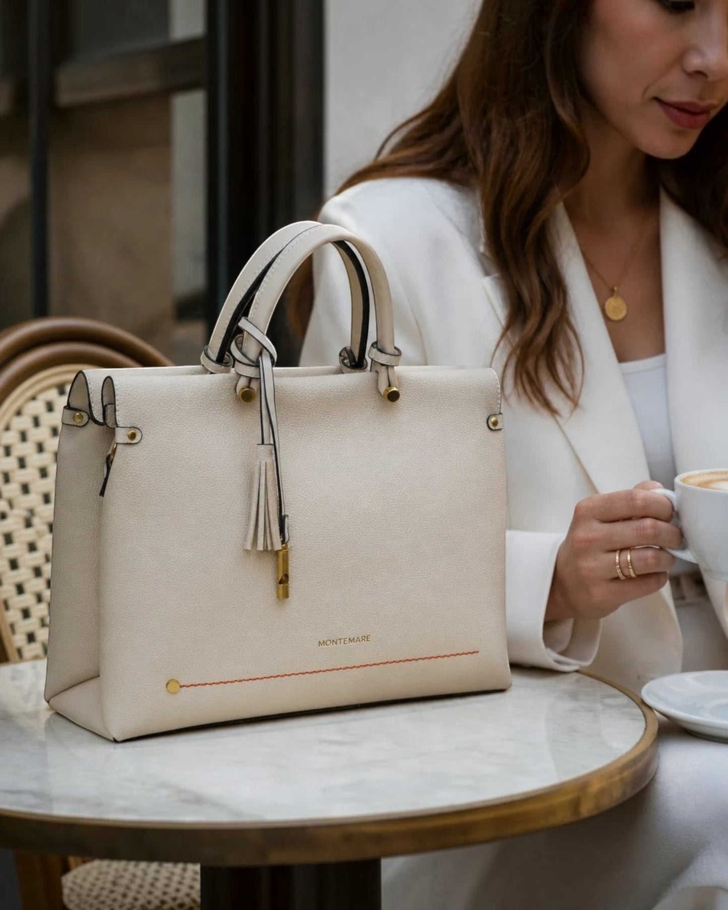 Montemaré Vienna small carry-on tote in cream vegan leather with a safety whistle tassel, sitting on a cafe table.