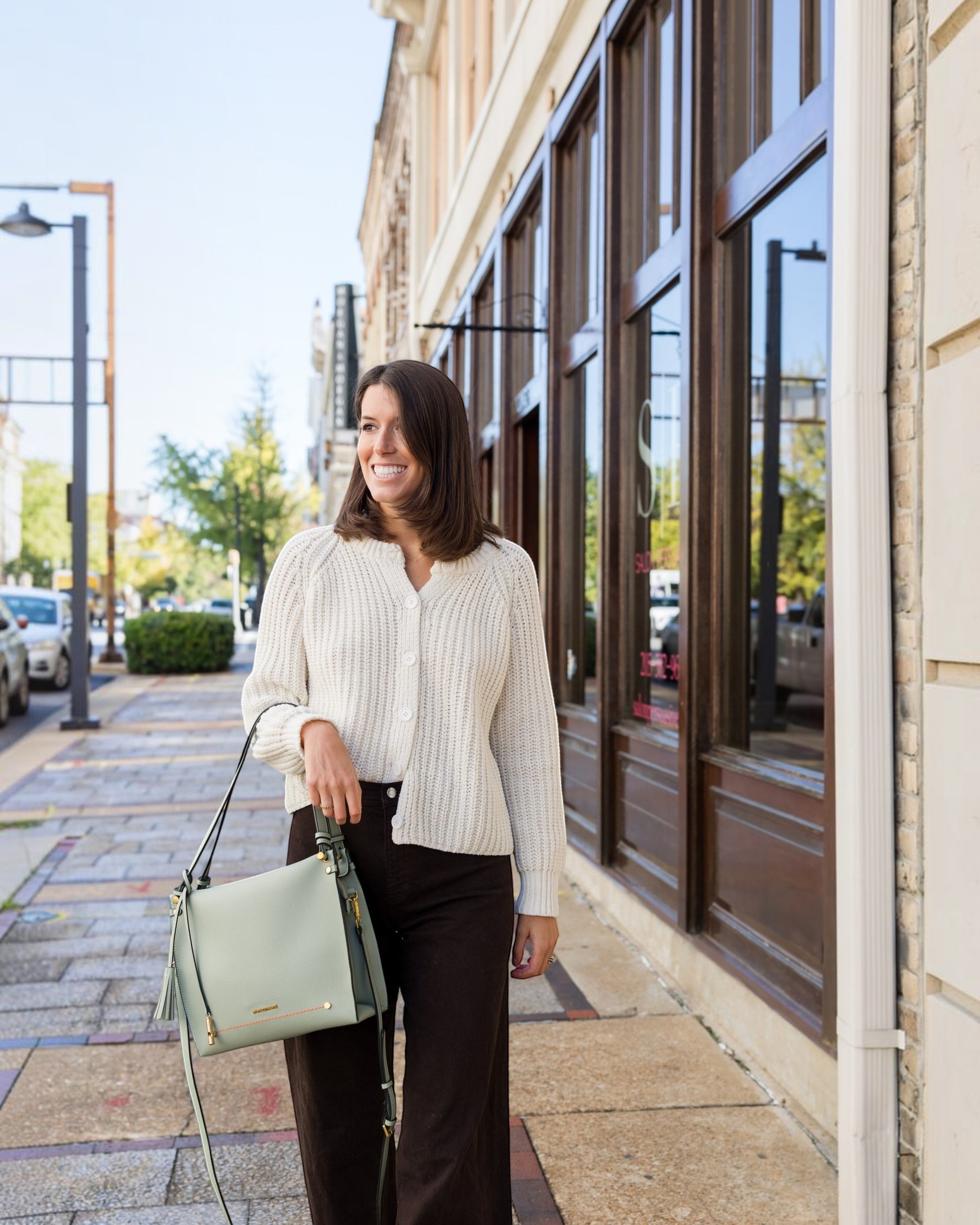 A woman wearing a Montemaré Vienna shoulder bag in sage green vegan leather while walking on a city sidewalk.