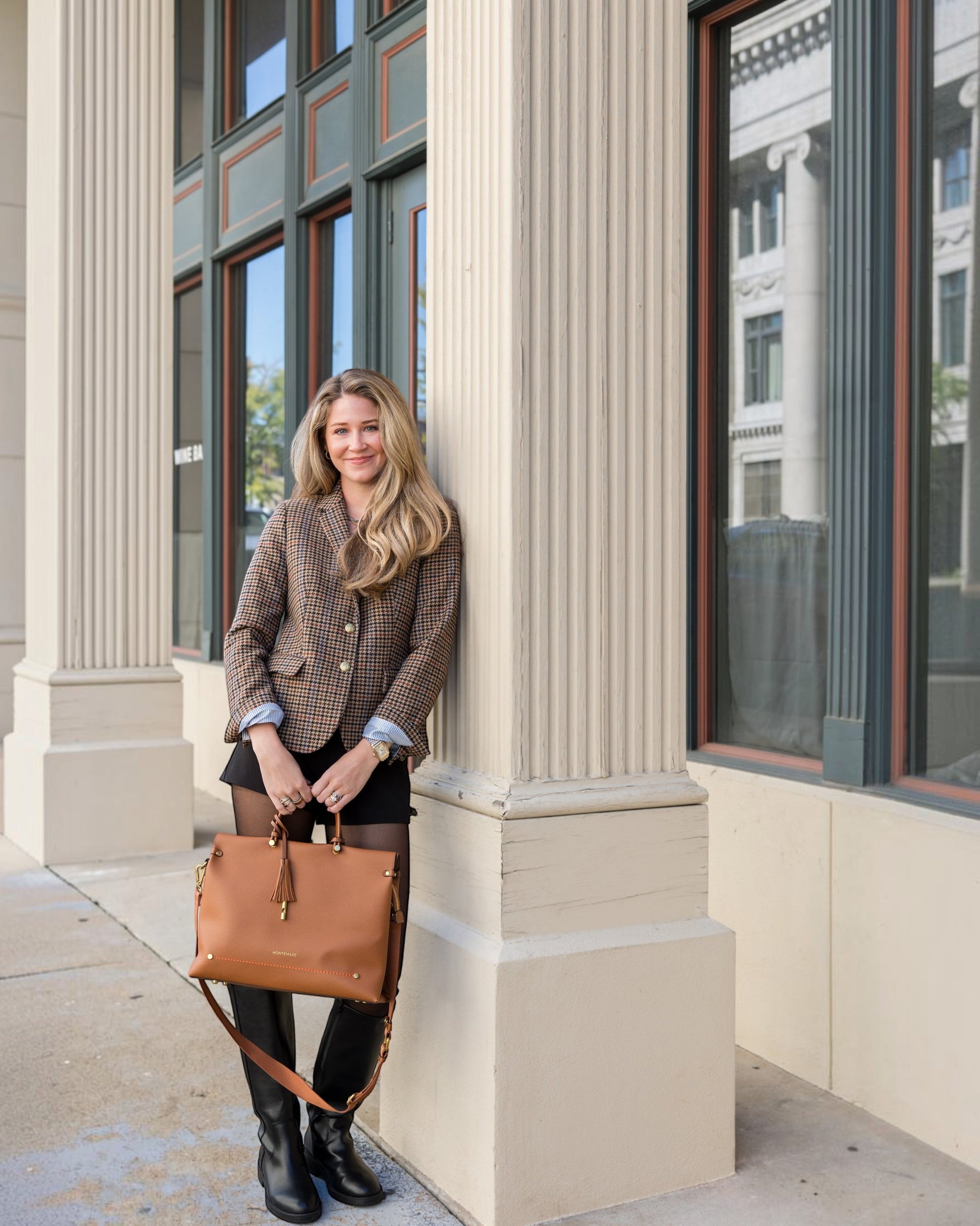 A woman holding a tan Montemaré Vienna vegan leather carry-on tote with a tassel detail while leaning against a column.