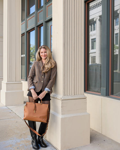 A woman holding a tan Montemaré Vienna vegan leather carry-on tote with a tassel detail while leaning against a column.